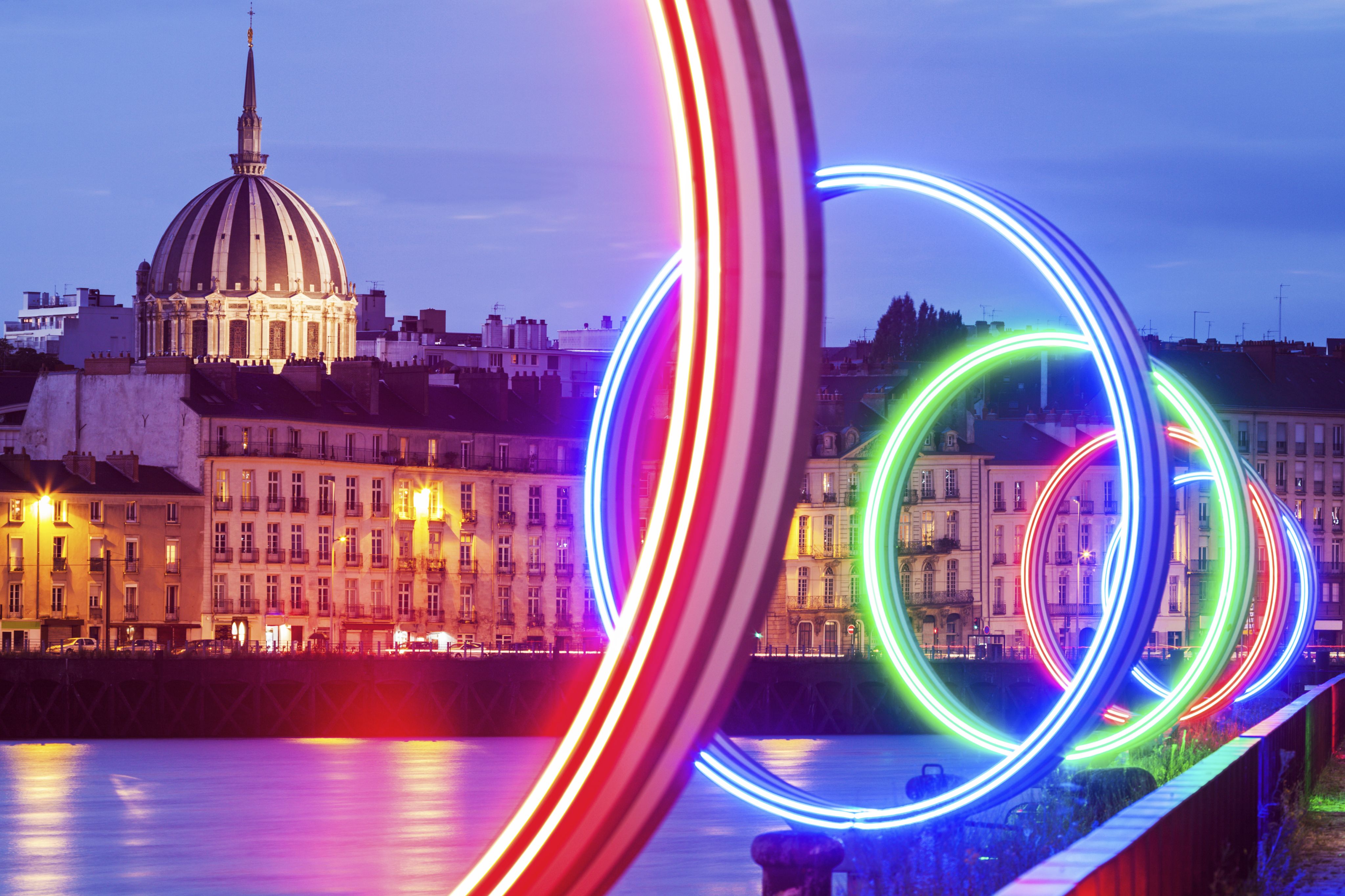 A bridge over the Loire river in Nantes, illuminated by circular neon lights, and a row of tall buildings with a dome in the background